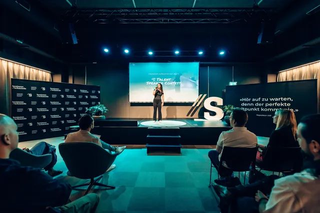 Shirin singt am Talent Speaker Contest der Business Schmiede, Bühne in blauem Licht, Publikum in Sesseln.